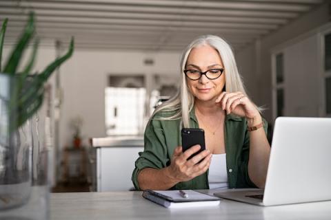 Woman looking at cell phone sitting by a laptop